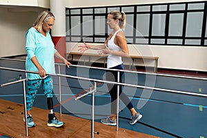 Female physiotherapist helping disabled senior woman walk with parallel bars in sports center