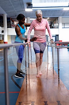 Female physiotherapist helping disabled senior woman walk with parallel bars in sports center