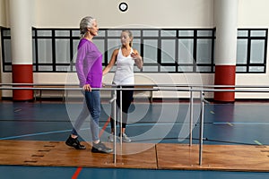 Female physiotherapist helping disabled senior woman walk with parallel bars in sports center