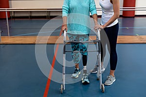 Female physiotherapist helping disabled senior woman walk with walker in sports center