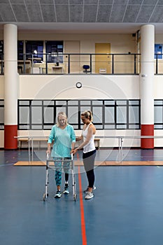Female physiotherapist helping disabled senior woman walk with walker in sports center