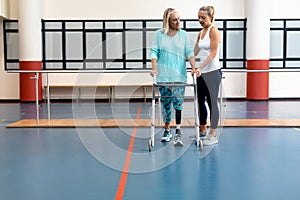 Female physiotherapist helping disabled senior woman walk with walker in sports center