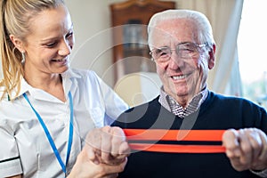 Female Physiotherapist Helping Senior Man To Use Resistance Band At Home