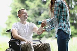 Female physiotherapist helps young man in wheelchair to lift weight of arms in park