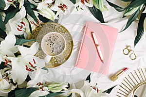Female pink notebook lies on white blanket with coffe cup, lipstick and many flowers. Flat lay, overhead view