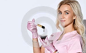 Female in pink scrubs and disposable gloves is posing isolated on white. Holding brush, glass bowl with black liquid. Close up