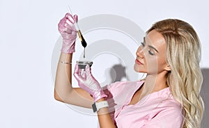 Female in pink scrubs and disposable gloves is posing isolated on white. Holding brush, glass bowl with black liquid. Close up
