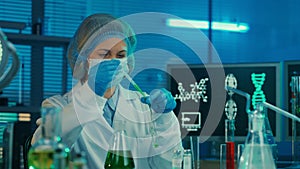 A female pipettes a green liquid into a glass test tube. A woman in a white medical gown, bonnet, blue gloves and a mask