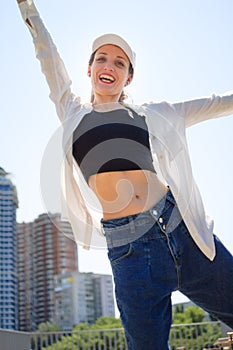 Female portrait of young active girl in black top, white shirt, basketball cap, and jeans on modern buildings background