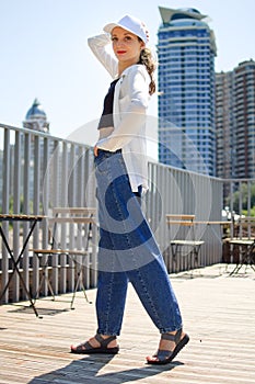 Female portrait of young active girl in black top, white shirt, basketball cap, and jeans on modern buildings background