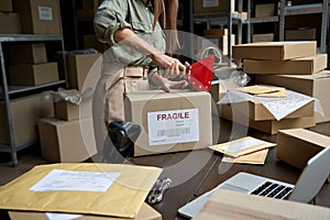 Female post mail storage worker holding tape dispenser sealing shipping box.