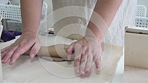 Female potter rolls out flatting piece of clay lying on fabric using wooden rolling pin in ceramic studio.