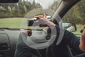 Female pouring the hot tea in tourist thermos mug. She sitting on co-driver seat inside modern car, enjoying the moody rainy day photo