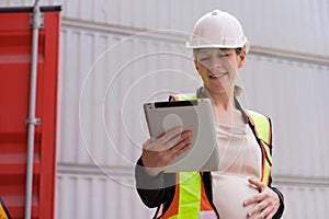 Female pregnant engineer or manager in safety helmet and vest holding digital tablet in front on container at cargo shipping yard