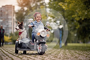 Female preschooler driving electrical motorcycle toy with sidecar and her dog in it, smiling