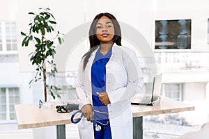 Female pretty African doctor in white coat looking to camera in an modern medical office