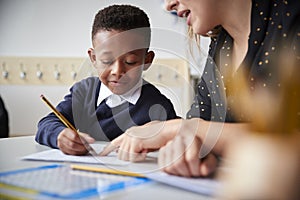 Female primary school teacher helping a young school boy sitting at table in a classroom, close up, selective focus