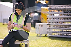 Female Production Worker With Mobile Phone Setting Up Outdoor Stage For Music Festival Or Concert