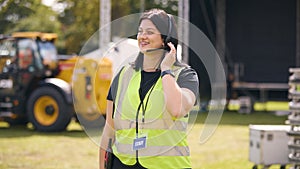 Female Production Worker Talking On Headset Setting Up Outdoor Stage For Music Festival Or Concert