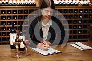 Female professional sommelier making notes on tablet sitting at the table and tasting red and white wine poured in