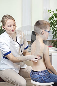 Female pulmonologist doctor during the medical examination. She listens to the boy`s lungs with a stethoscope in the doctor`s offi photo