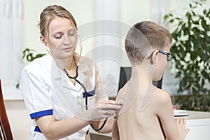 Female pulmonologist doctor during the medical examination. She listens to the boy`s lungs with a stethoscope in the doctor`s offi