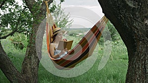 Female relaxing in hammock at green garden. Caucasian woman lying in hammock, reading book and resting at countryside. Girl in hat