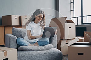 Female renter homeowner sitting on armchair with laptop orders removal service on moving day