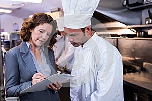 Female restaurant manager writing on clipboard while interacting to head chef