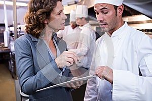 Female restaurant manager writing on clipboard while interacting to head chef