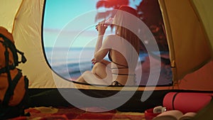 Female resting on campsite relaxing near ocean. Young woman sitting outside the tent at sunset on the beach drinking