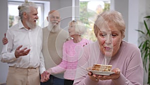 Female retiree smiling blowing out candle making wish on birthday looking back at blurred friends clapping rejoicing