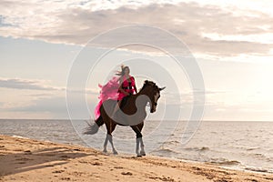 Female rider wearing a pink dress galloping along a beach on a brown horse.