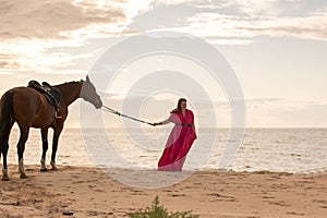 Female rider wearing a pink dress galloping along a beach on a brown horse.