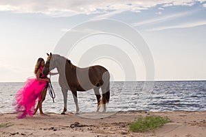 Female rider wearing a pink dress galloping along a beach on a brown horse.