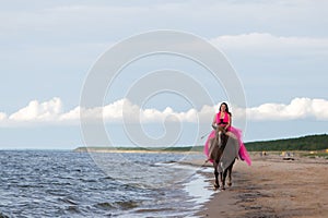 Female rider wearing a pink dress galloping along a beach on a brown horse.