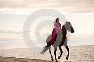 Female rider wearing a pink dress galloping along a beach on a brown horse.