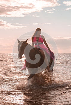 Female rider wearing a pink dress galloping along a beach on a brown horse.