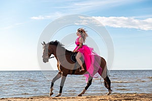 Female rider wearing a pink dress galloping along a beach on a brown horse.