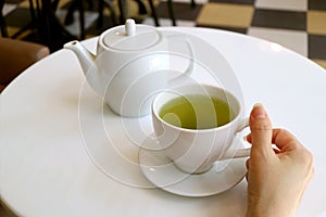 Female`s hand holding cup of green tea on a white round table with teapot in background