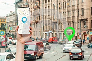 A female's hand holds a smartphone with an online map app. In the background is a blurred city street with traffic