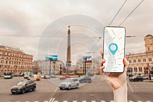 A female's hand holds a smartphone with an online map app. In the background is a city road with traffic. Concept of