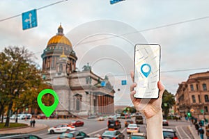 A female`s hand holds a smartphone with an online map app. In the background is a city street with building and green location