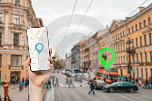 A female`s hand holds a smartphone with an online map app. In the background is a city street with traffic, with a green location