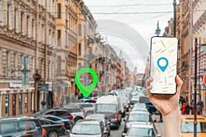 A female's hand holds a smartphone with an online map app. In the background is a city street with traffic jam, with