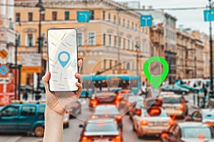 A female`s hand holds a smartphone with an online map app. In the background is a city street with traffic jam and green location