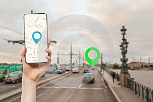 A female`s hand holds a smartphone with an online map app. In the background is a road with car and green location icon.Concept o