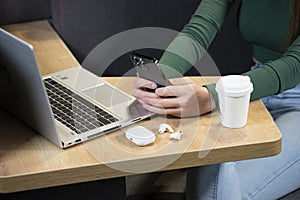 Female`s hands with cell phone in front of laptop computer, sits at cafeteria, surrounded with mug of coffee and modern laptop photo