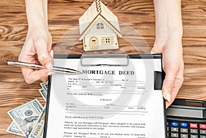 Female's hands holding clipboard with mortgage deed and pen over table with money, calculator and model of house