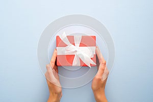 Female`s hands holding red gift box with white ribbon bow on pastel blue background. Christmas, New Year, Valentine`s day and